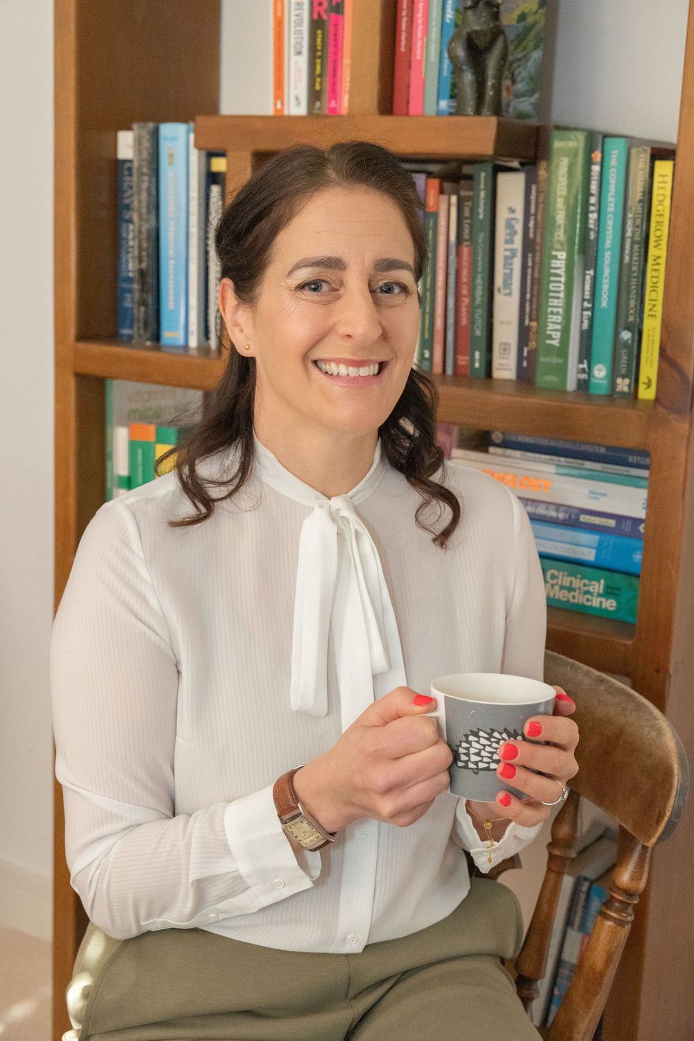 Person in white blouse sits in front of bookshelf with wooden chair enjoying coffee or tea from small cup.