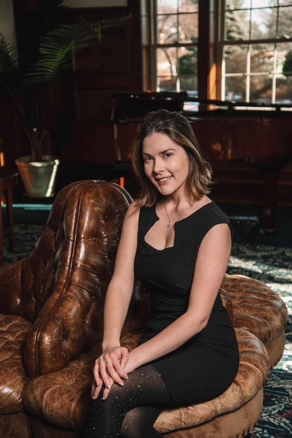 Smiling young woman in black dress sits relaxed on tufted leather chair in warmly lit lounge.