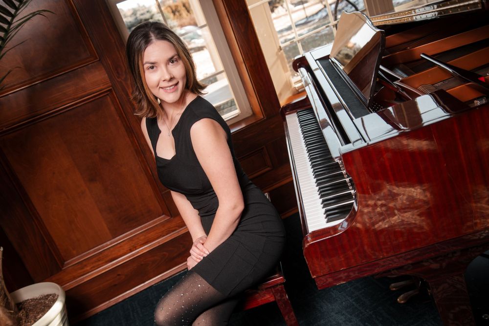 Smiling brunette woman in black dress sits beside a polished grand piano in elegant room.