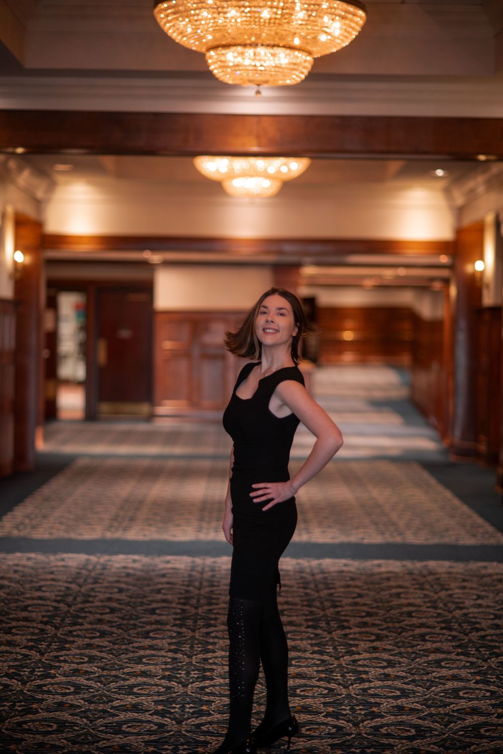 A woman in a black dress and boots stands confidently in an elegant chandelier-lit hallway.