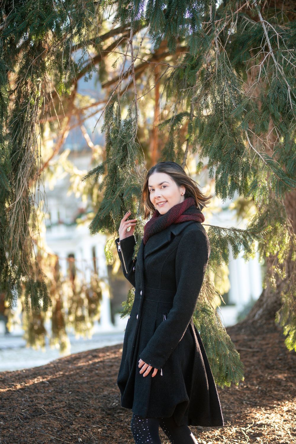 A smiling woman in a black coat and red scarf stands under a pine tree outdoors.