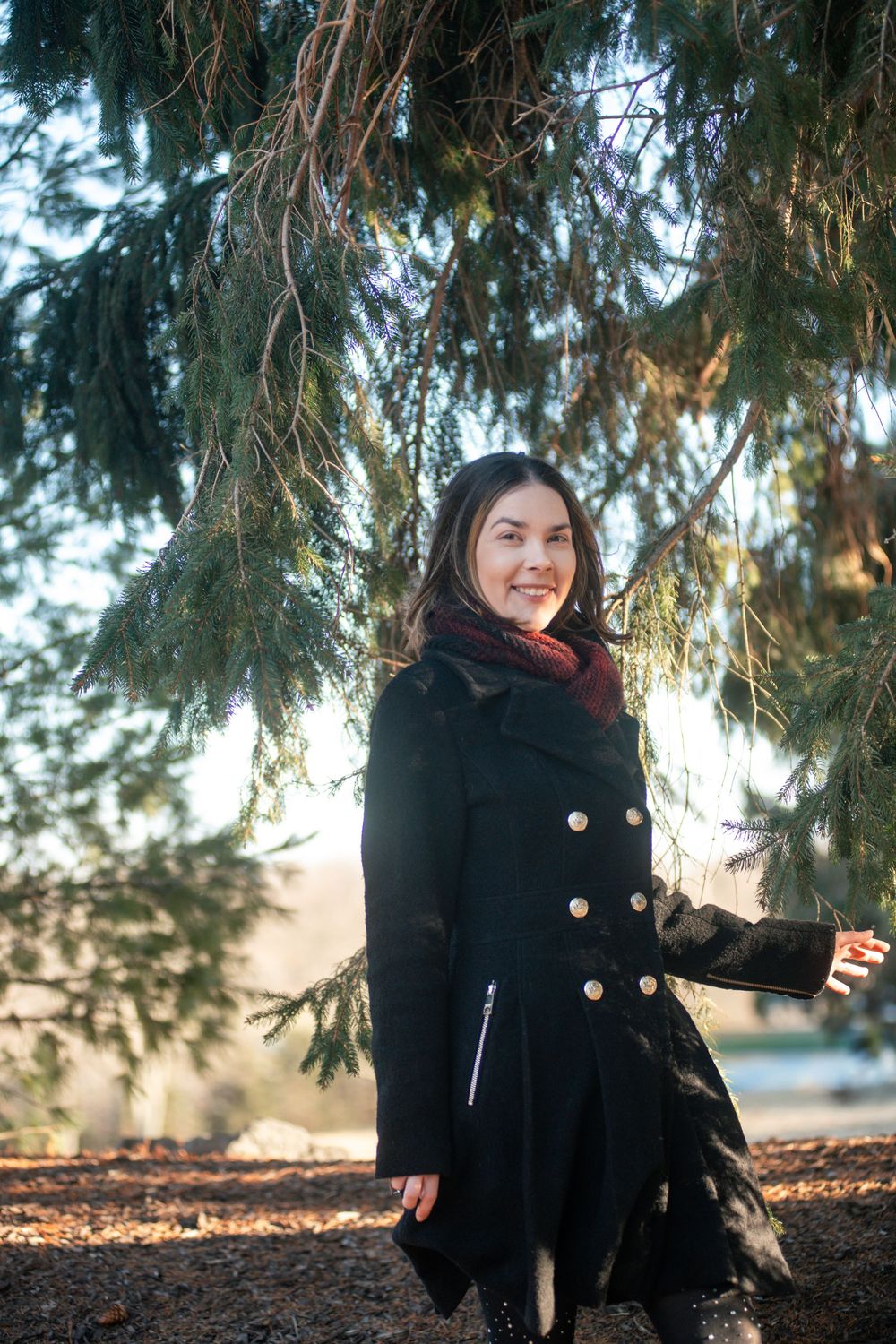 A smiling young woman in a black coat with silver buttons stands under pine tree branches.