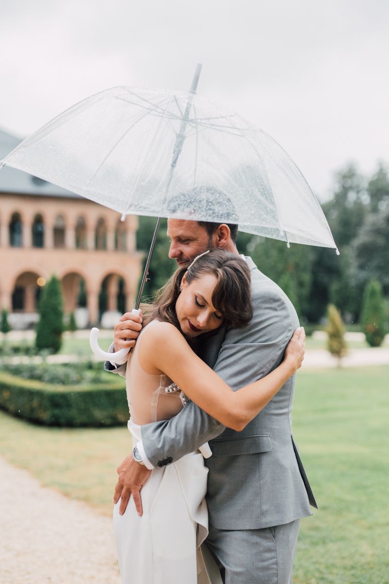 A couple shares an intimate embrace under a clear umbrella on a rainy day at an outdoor venue.
