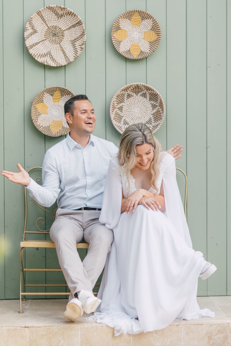A couple in white outfits shares joyful moments against a sage green wall decorated with woven basket wall art.