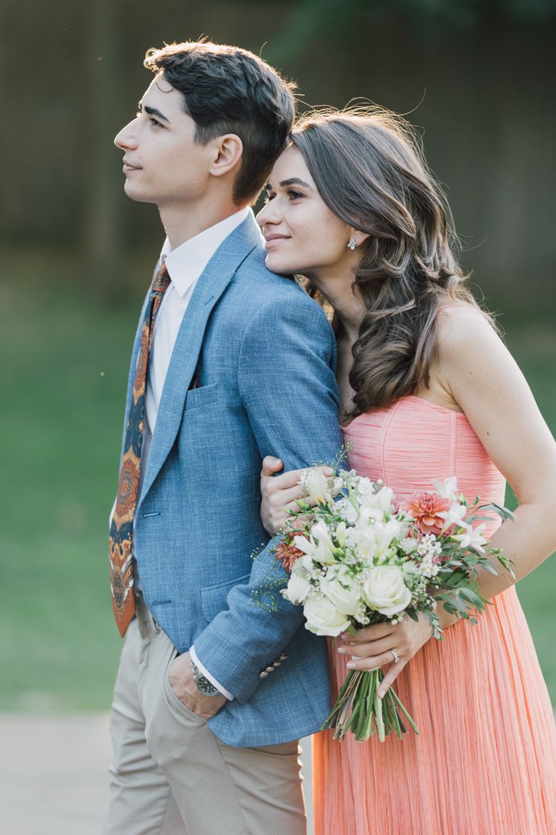 A couple in formal attire pose together outdoors, with the woman holding a bouquet of white and coral flowers.