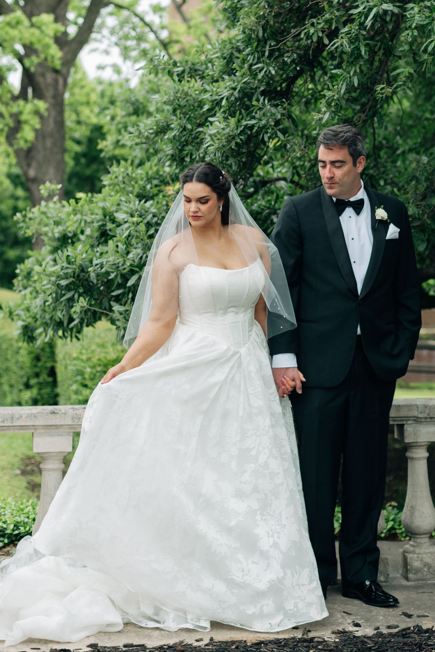 Bride in white lace gown and veil holds hands with groom in black tuxedo in lush garden setting.