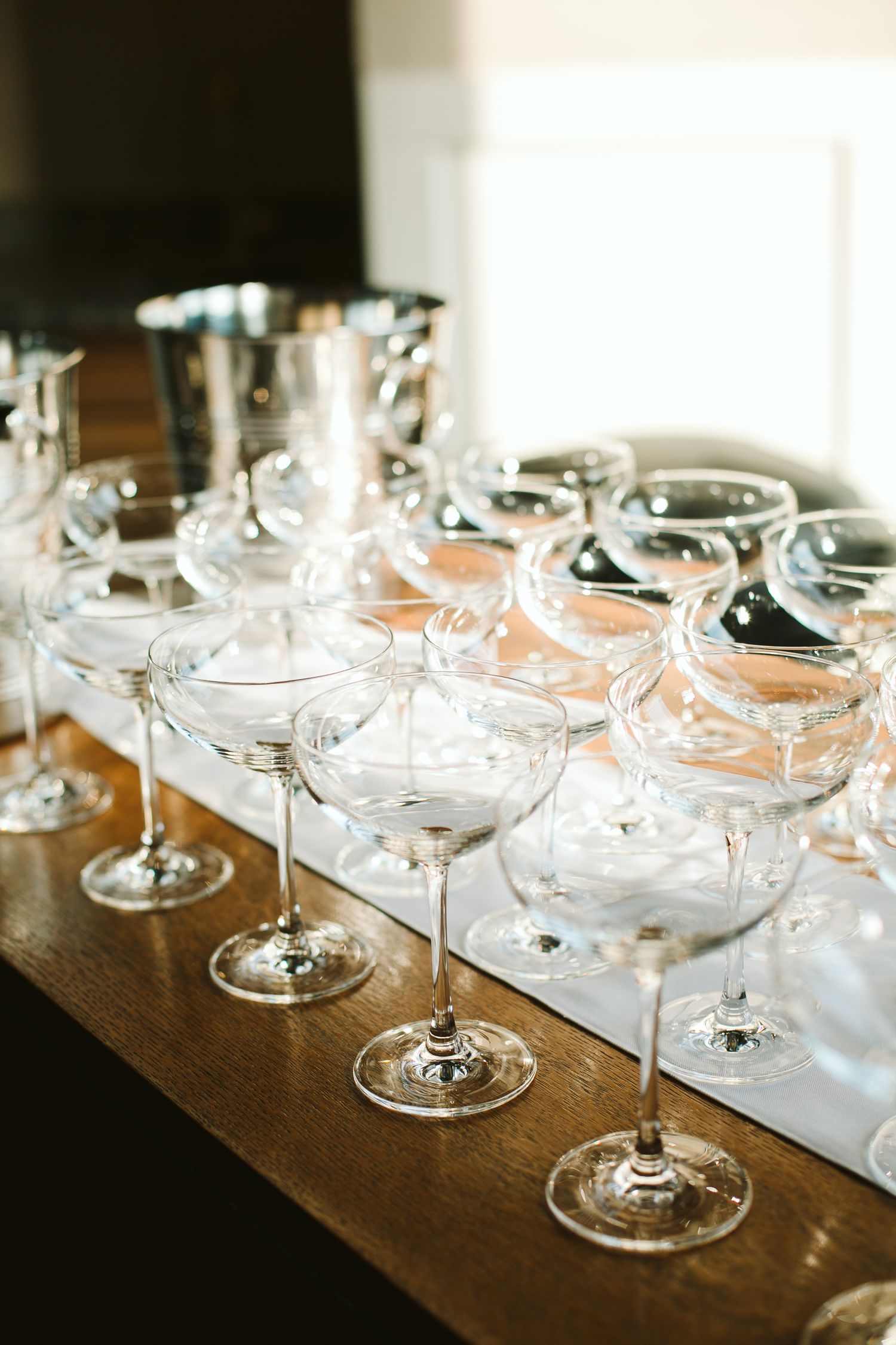 Rows of empty crystal coupe champagne glasses arranged on a wooden bar table for an elegant event.