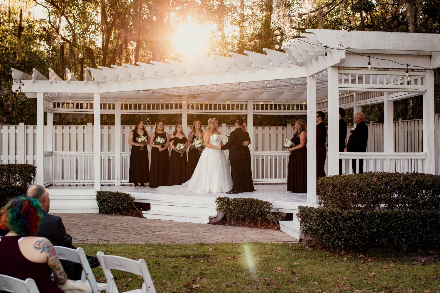 Outdoor wedding ceremony takes place at sunset under a classic white wooden gazebo with decorative details.