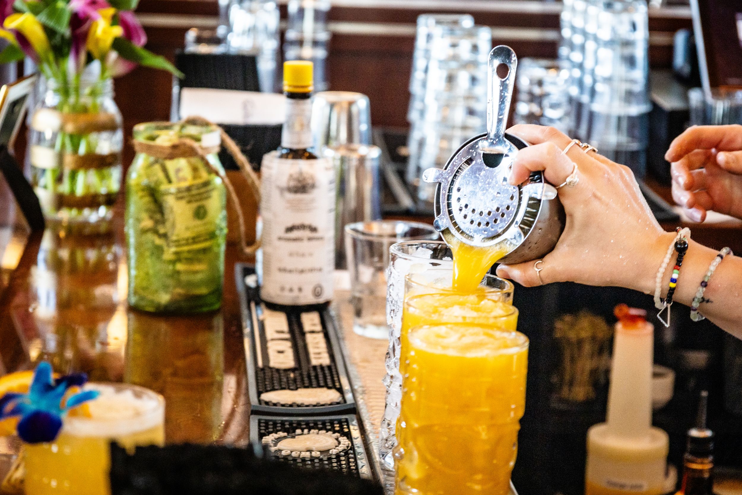 Bartender pouring bright yellow cocktail into glass with various bottles and bar equipment in background.