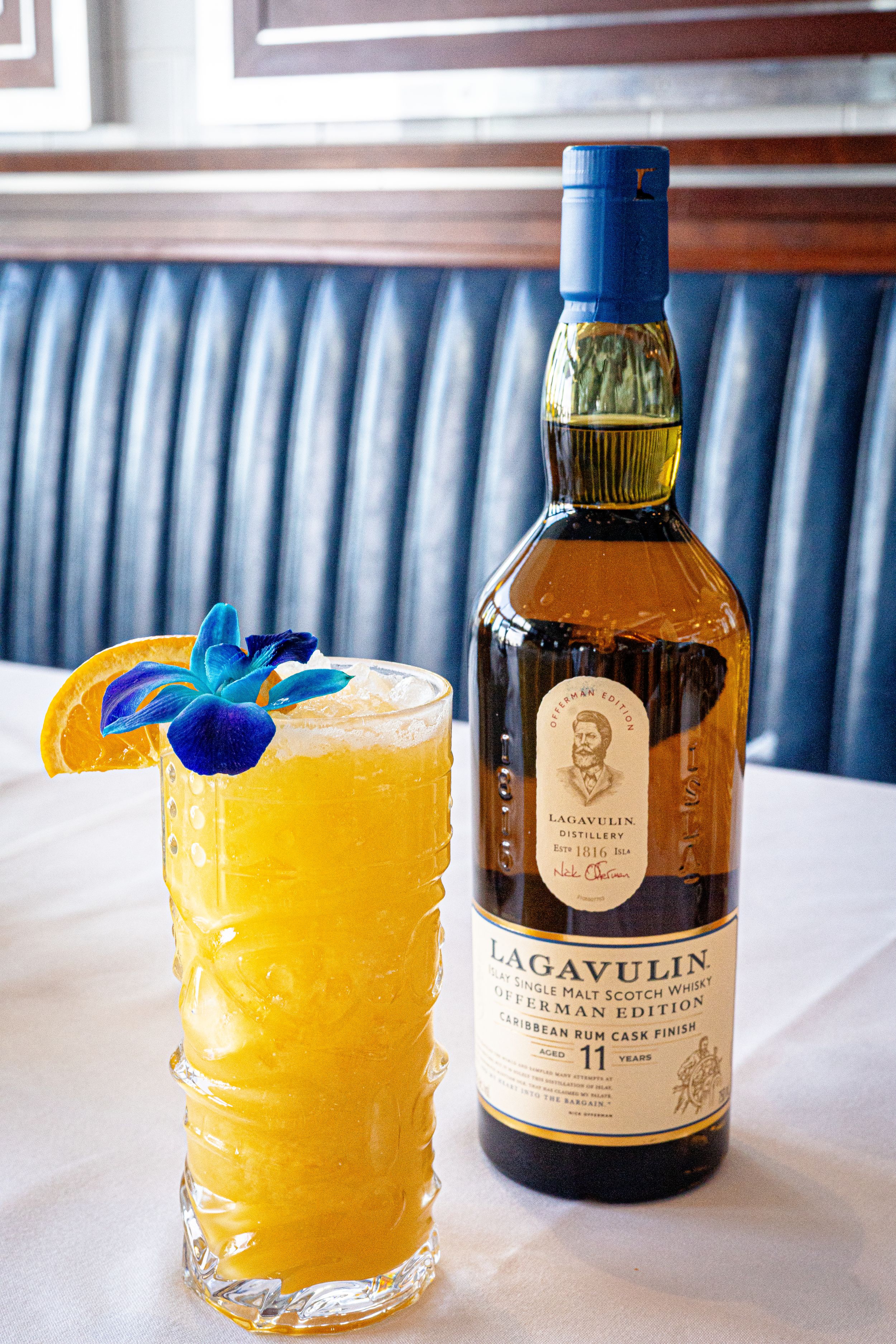Lagavulin whisky bottle next to garnished yellow cocktail with blue flower on white tablecloth.