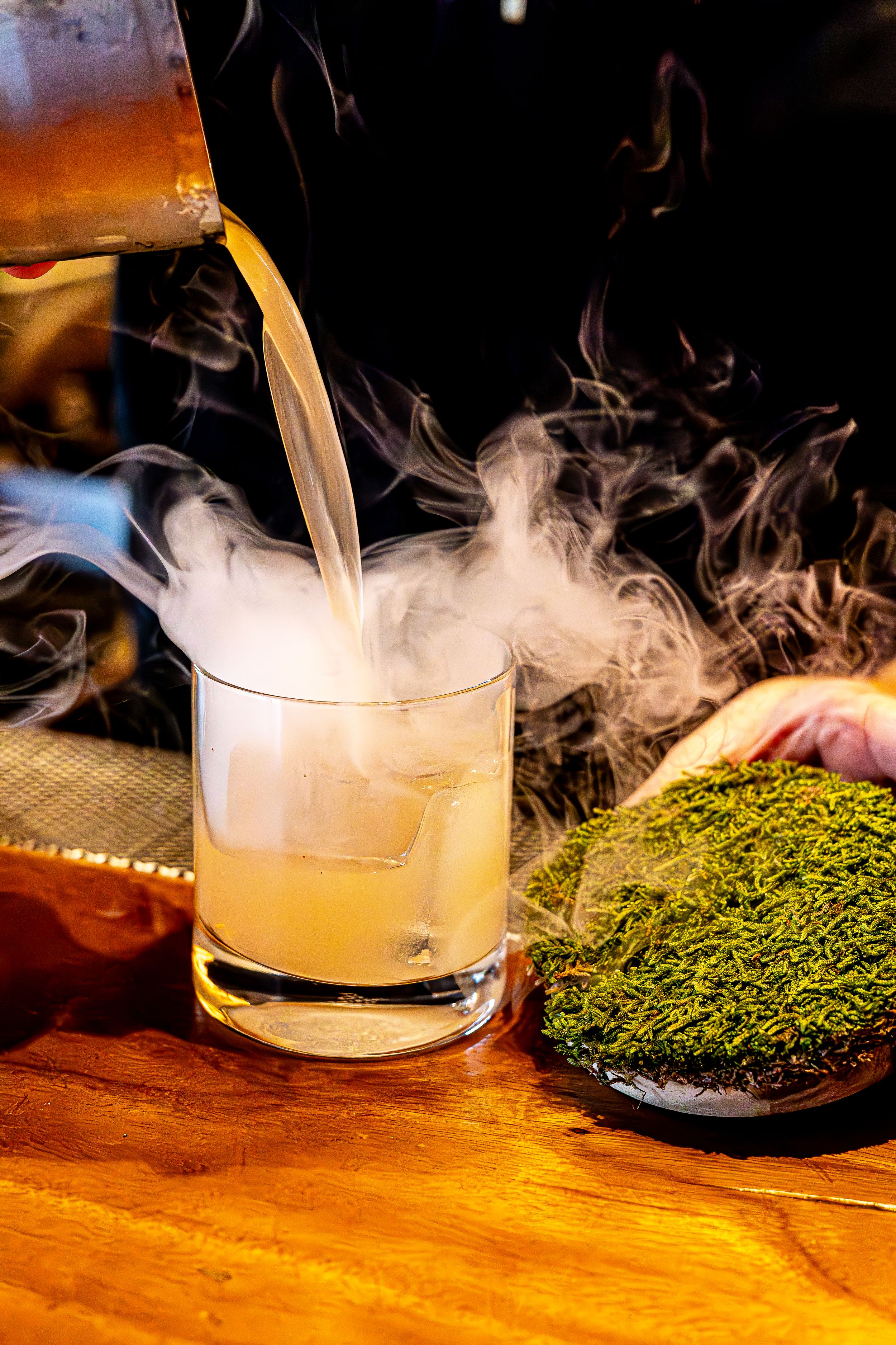 Smoky cocktail being poured next to a green-topped dessert on wooden bar surface.