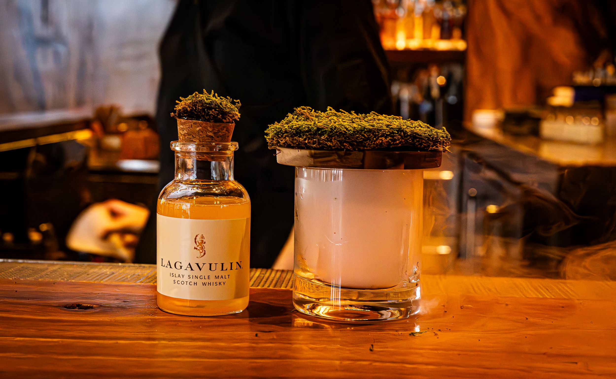 Craft cocktail with moss garnish served alongside Lagavulin bottle on wooden bar.