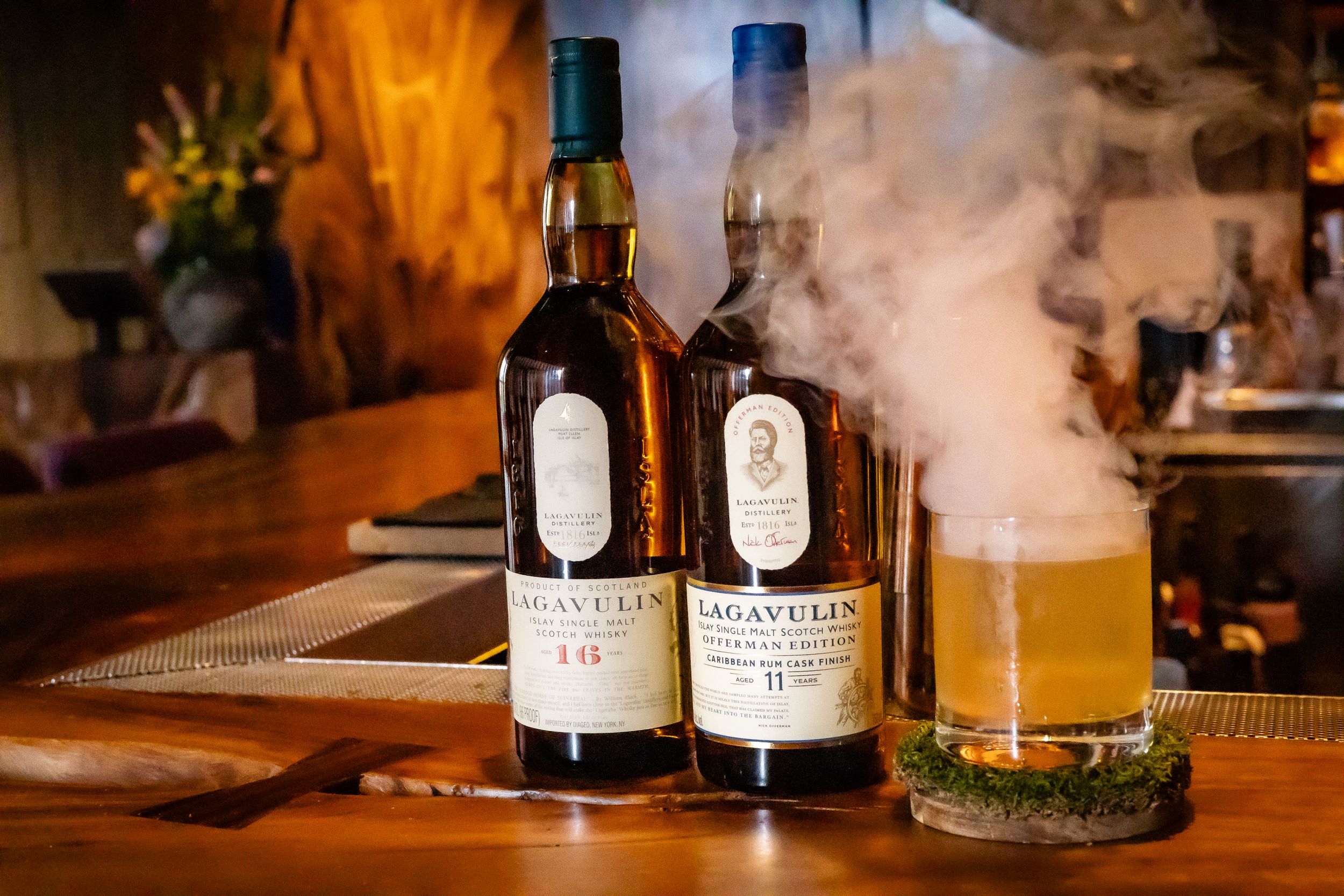 Two bottles of Lagavulin whisky displayed with smoking cocktail on bar counter.