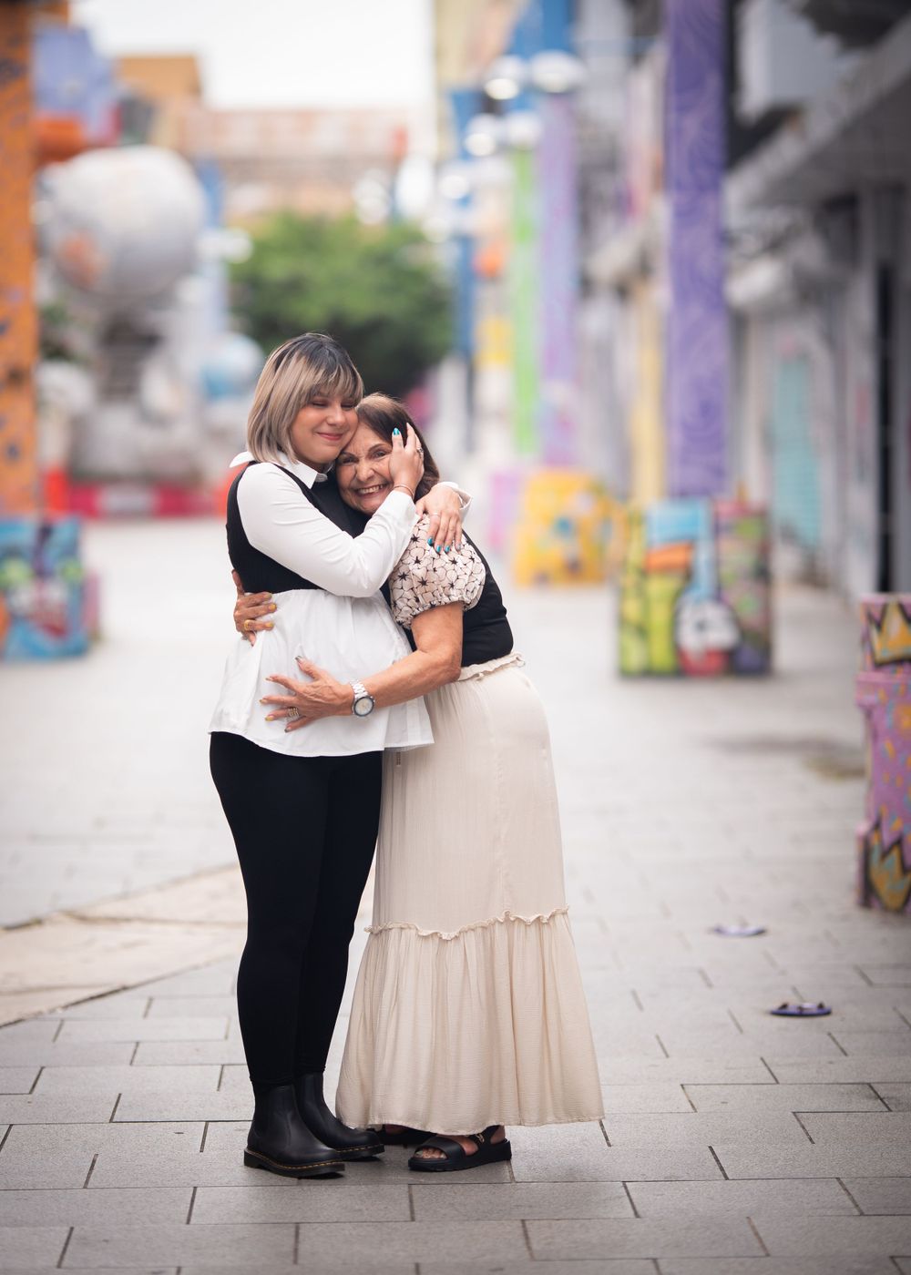 Two people embrace warmly on a colorful street with graffiti murals in the background.
