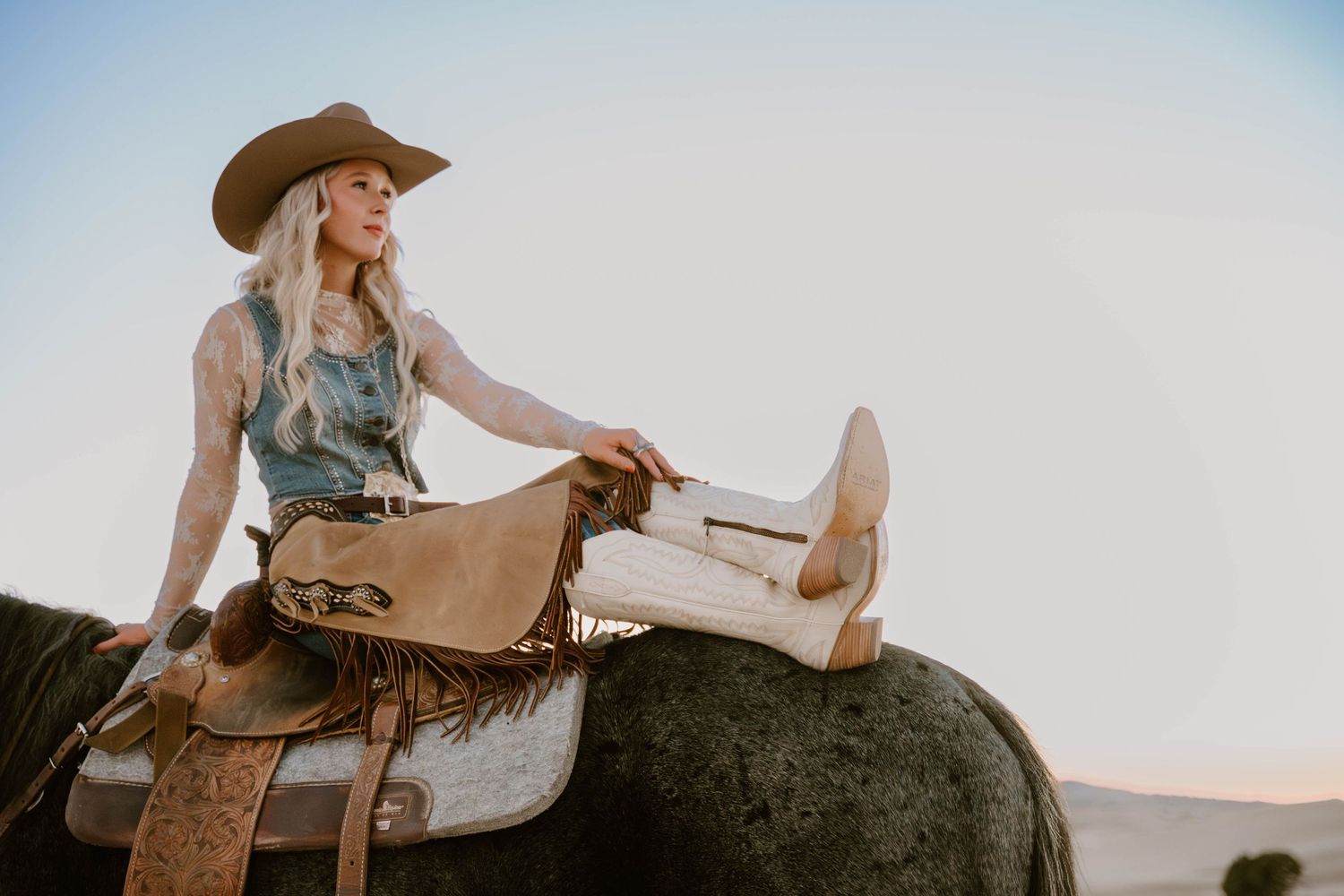 Cowgirl in chaps and Ariat Boots relaxing on horseback against bright sky background.