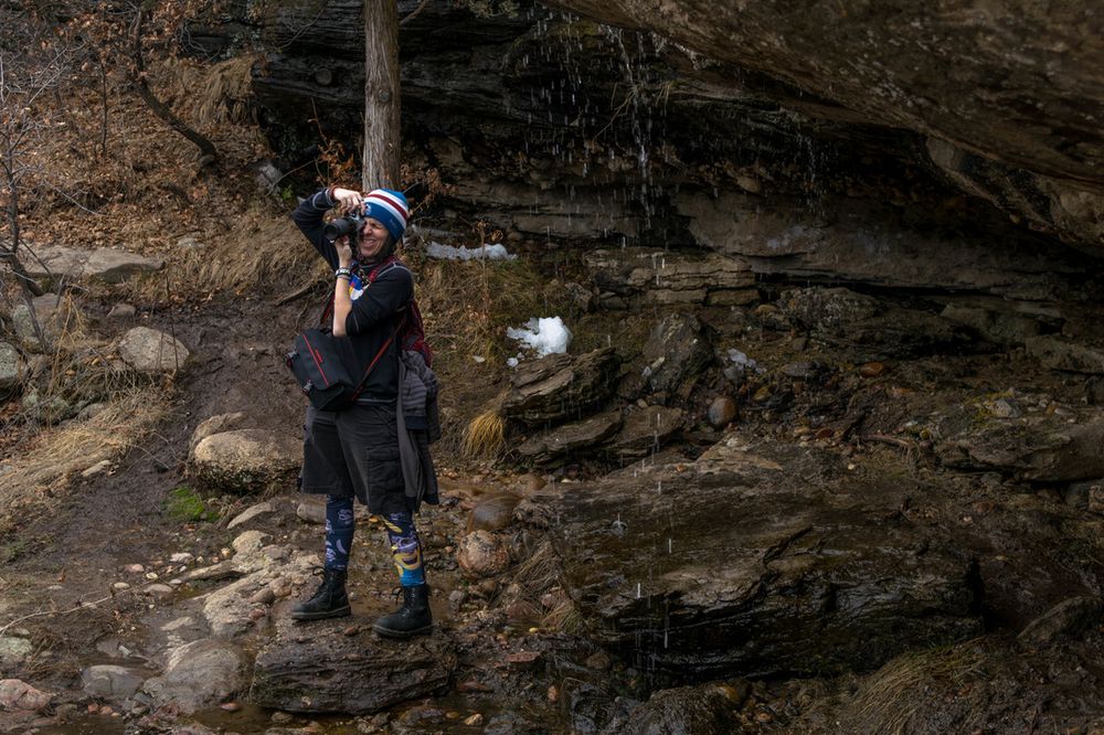A photographer stands beneath a rocky overhang near a waterfall.