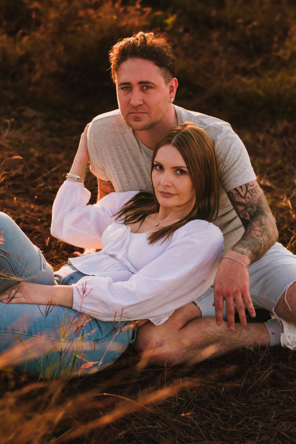 Romantic couple sitting in golden grass during sunset wearing white and casual attire.