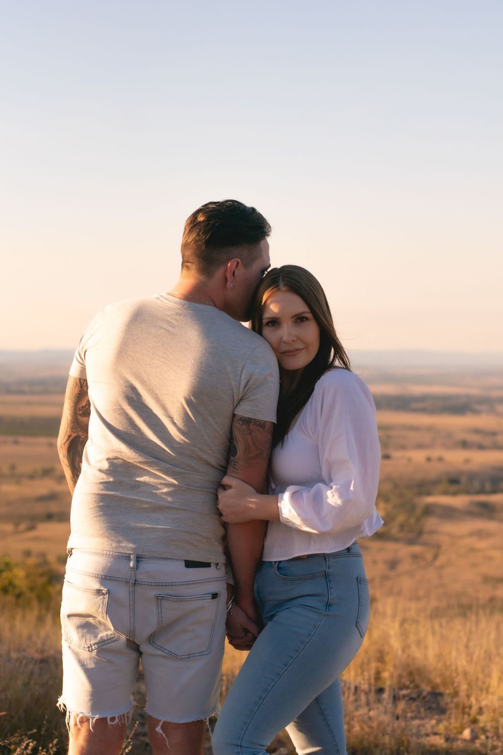 Couple in casual wear embracing on hilltop during sunset.