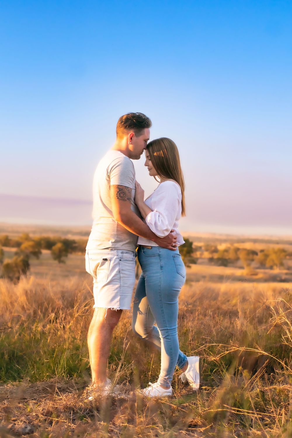 Couple standing close in golden field with blue sky background.