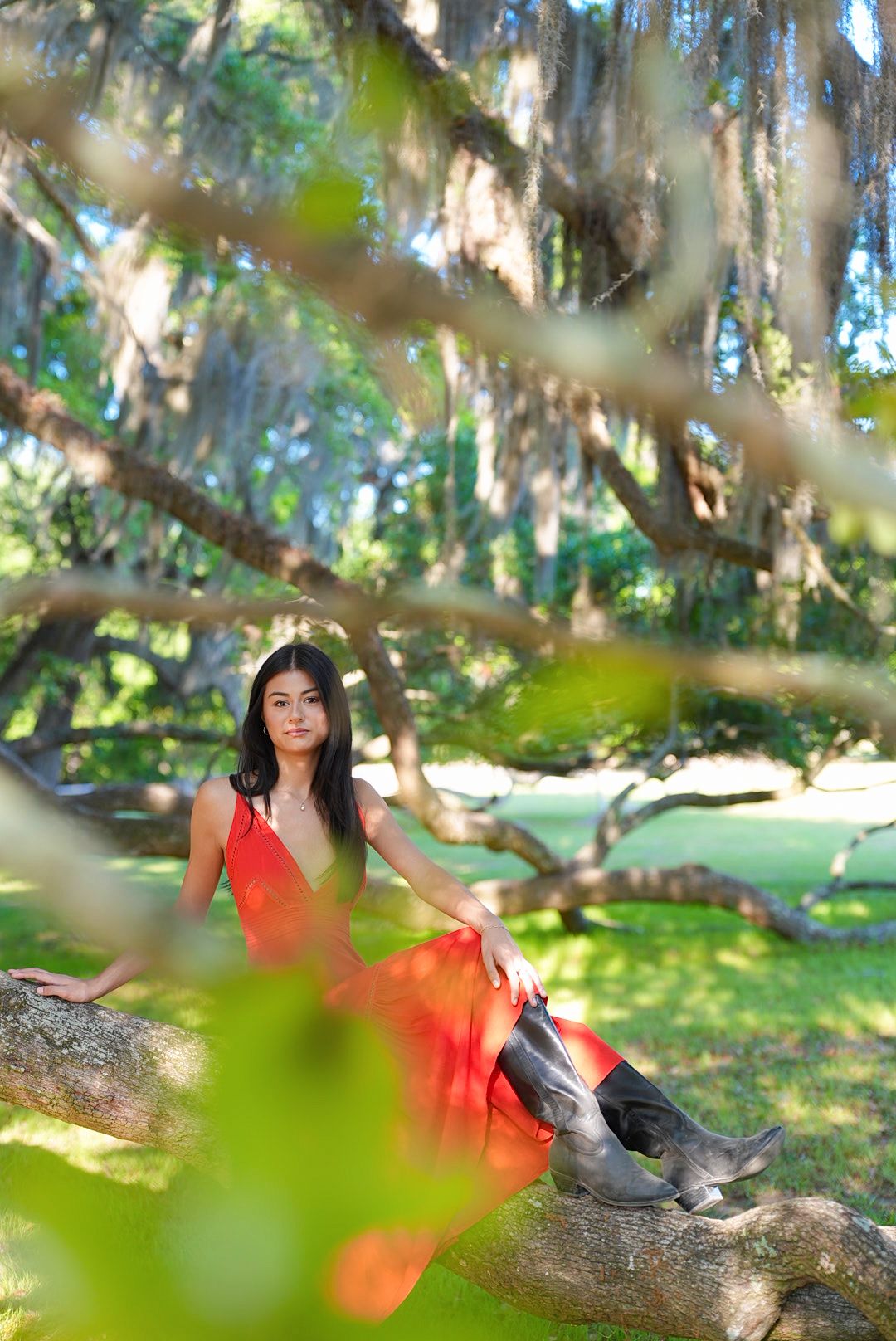 A woman in a red dress and tall boots sits on a thick tree branch beneath Spanish moss in a sunlit green park.