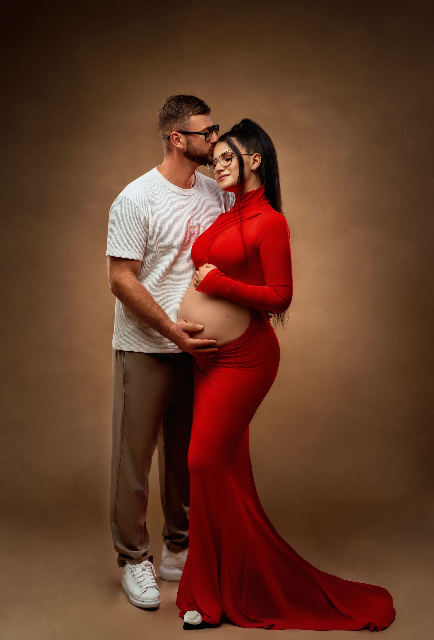 A couple embraces tenderly during a maternity photoshoot, with the mother wearing an elegant red flowing dress.