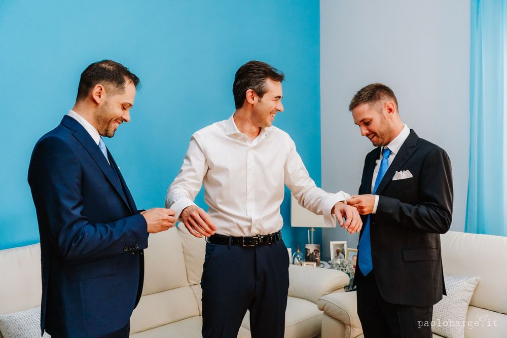 Three men in formal suits sharing a lighthearted moment against a bright blue wall in a living room setting.
