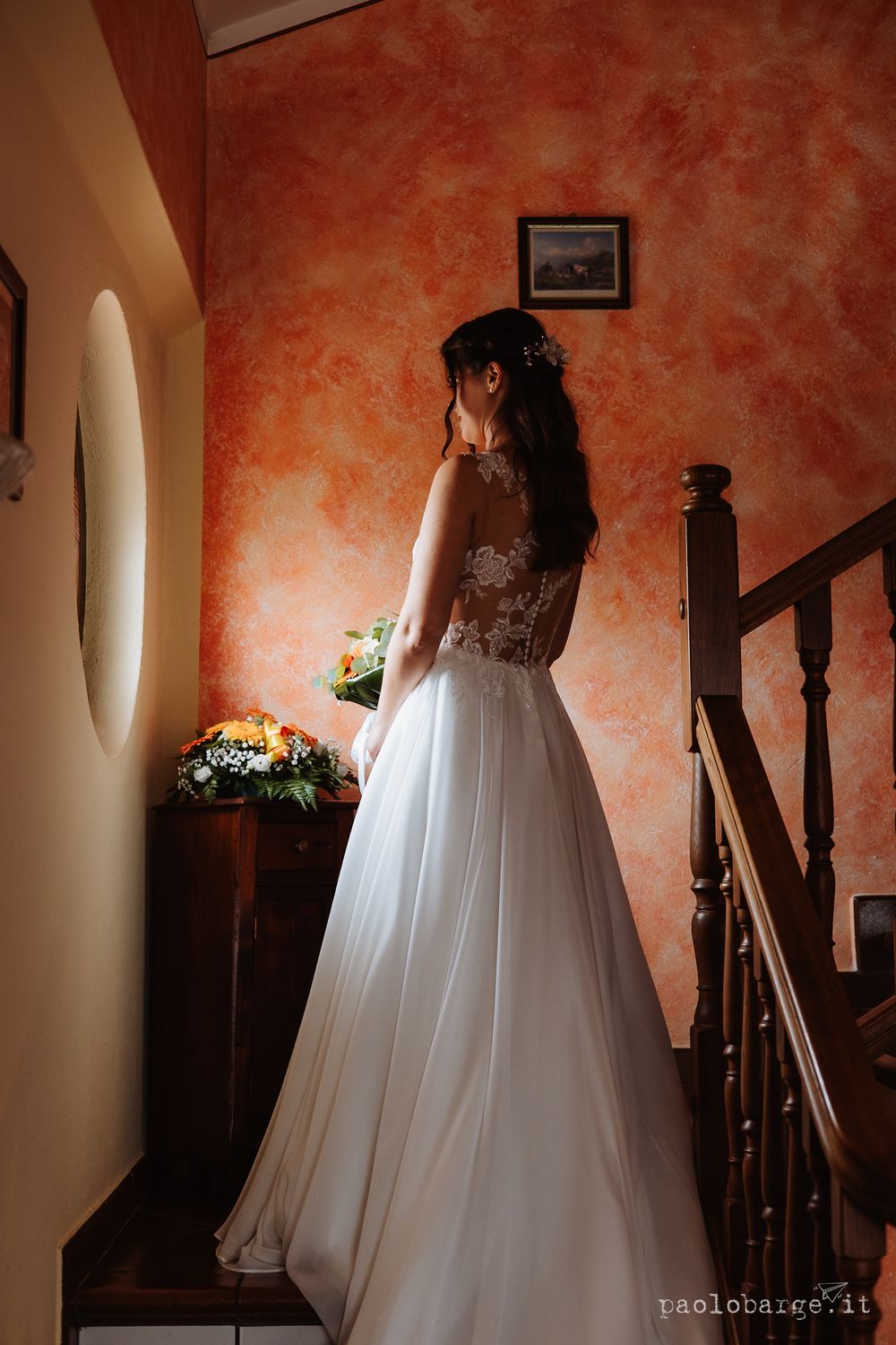 Bride in an elegant lace wedding gown standing on stairs against a textured orange wall.