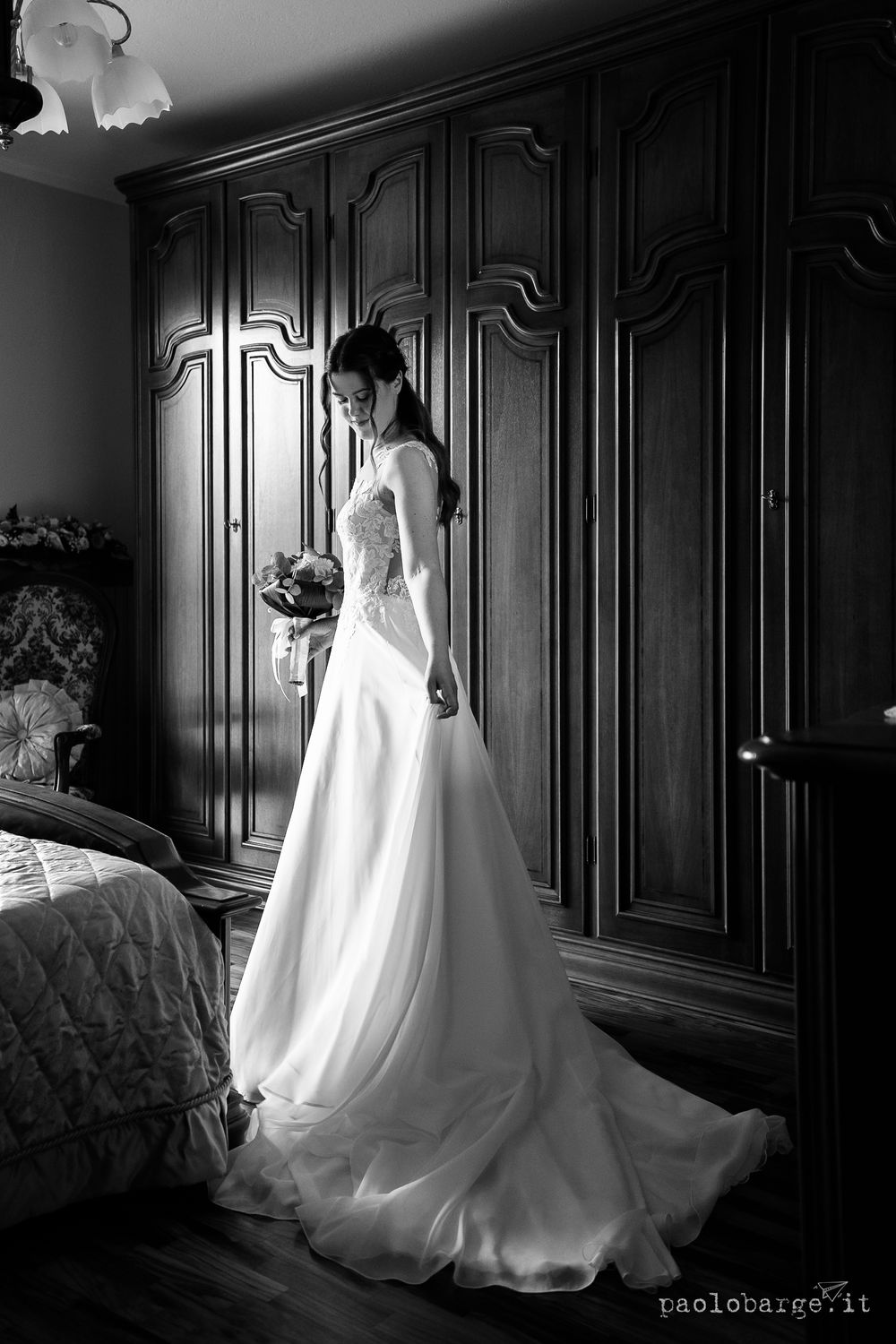 Dramatic black and white portrait of a bride in a flowing wedding gown beside ornate wardrobes.