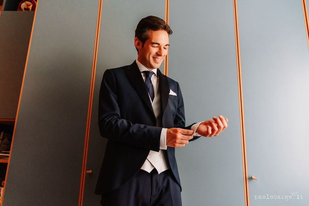 Man in navy suit adjusting his cufflinks against an orange-tinted wall.
