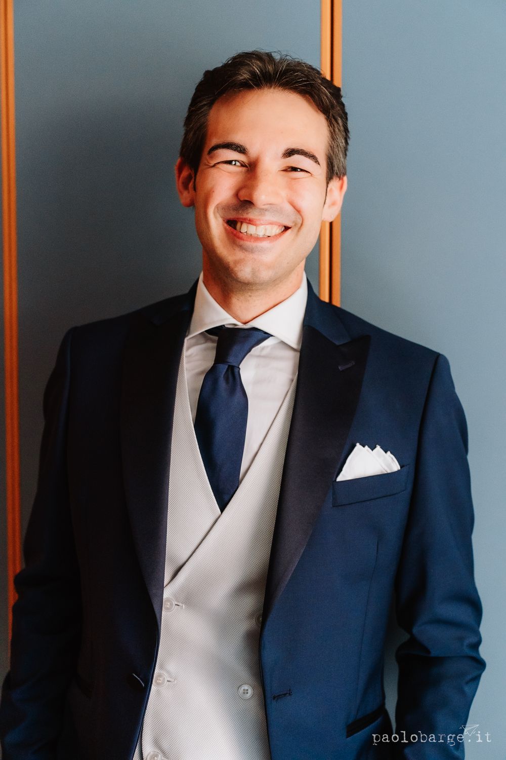 A groom smiles joyfully while wearing a navy suit with light gray vest and white pocket square.
