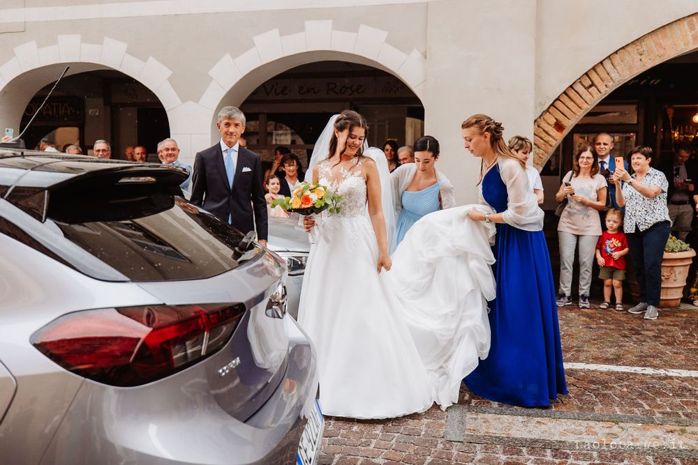 A bride in white dress exits with guests near a car under stone archways.