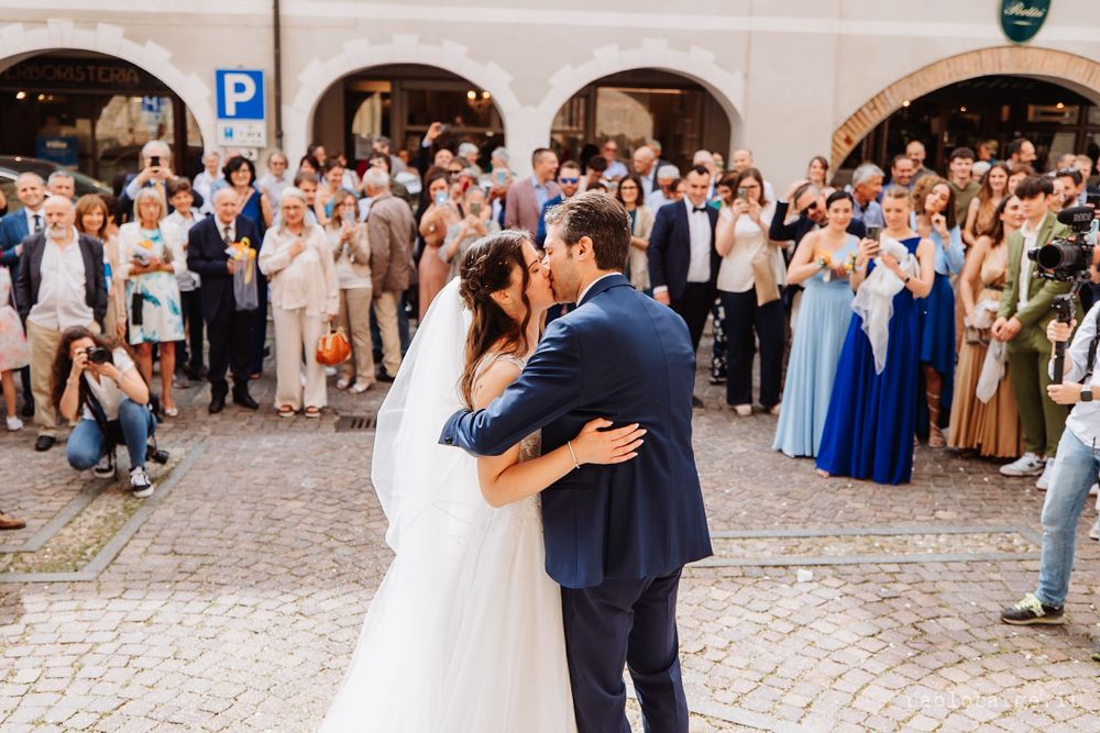 Wedding couple sharing first kiss while guests celebrate in historic courtyard.