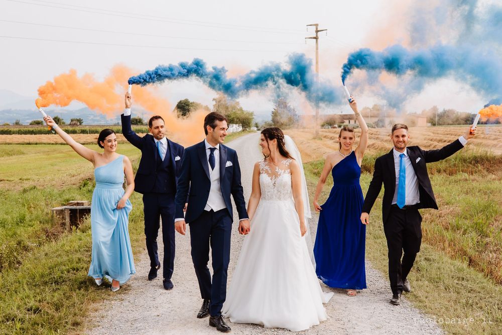 Bridal party celebrating with orange and blue smoke bombs in rural setting.