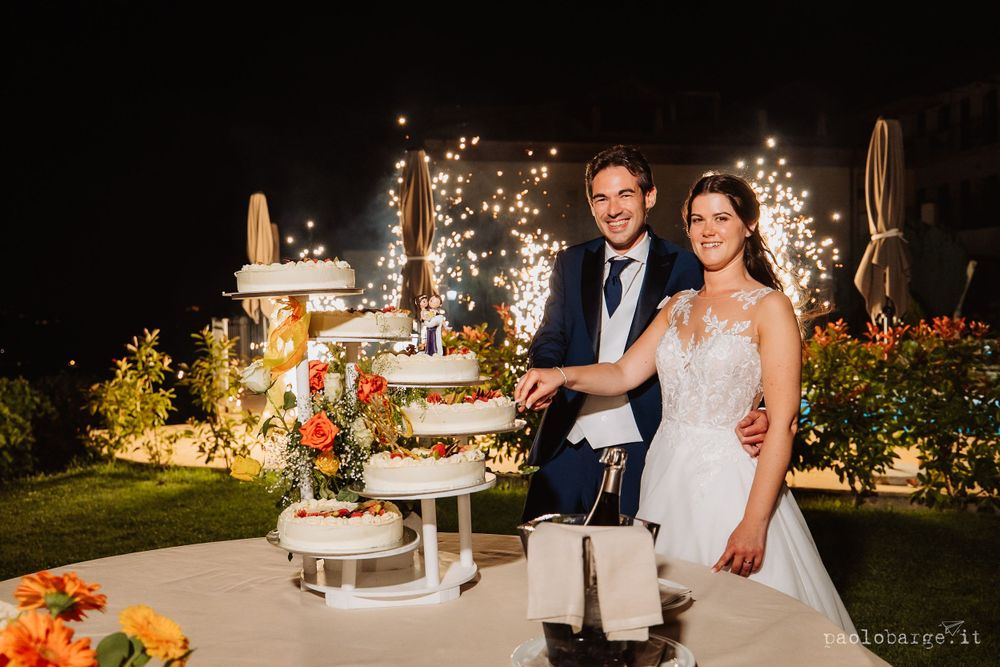 Couple cuts elegant tiered wedding cake at night with twinkle lights backdrop.