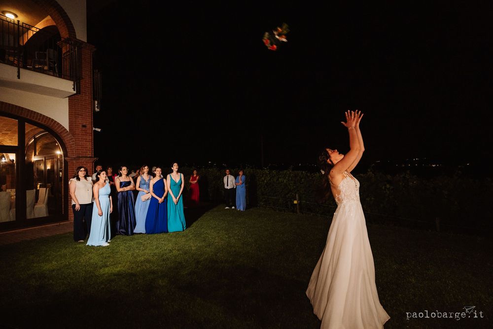 Wedding guests in blue dresses gather on a lawn at night while a bouquet is tossed into the air.