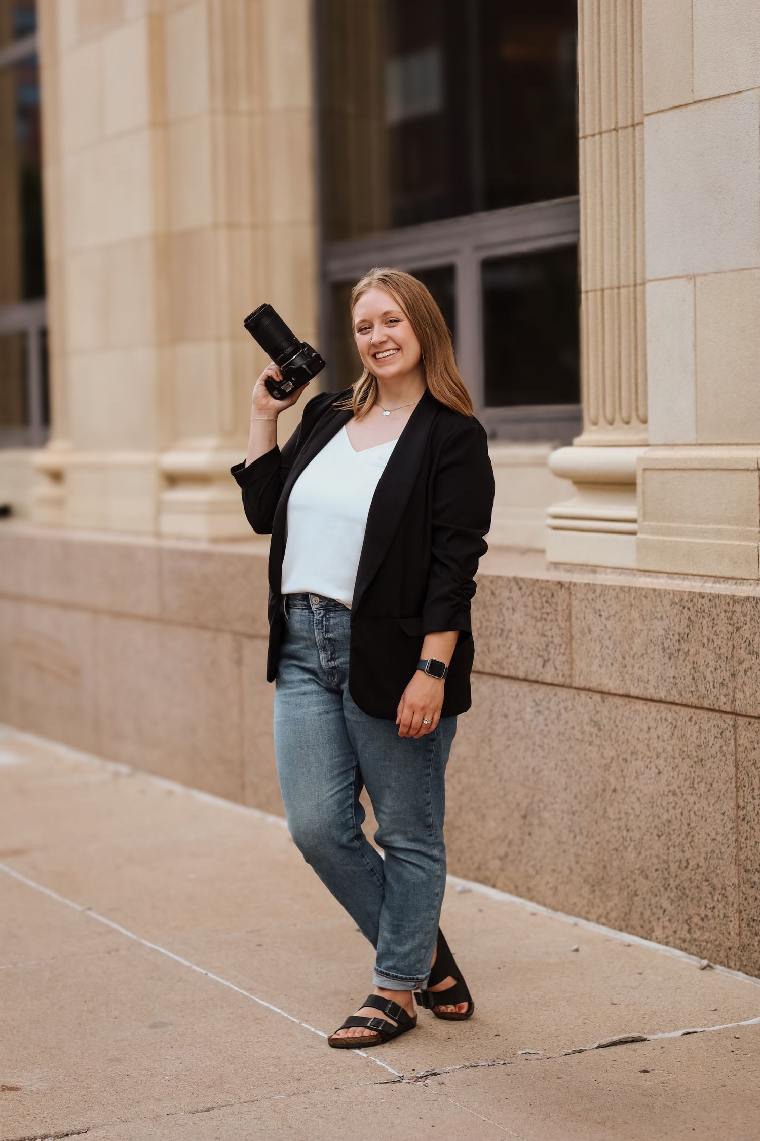 A portrait of a blonde woman holding a camera and smiling in front of a tan building