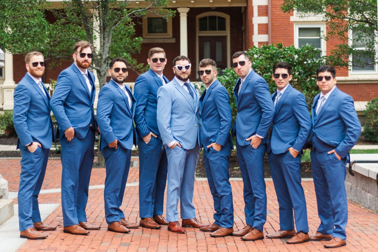 Groomsmen wearing sunglasses and blue suits pose together on brick steps.