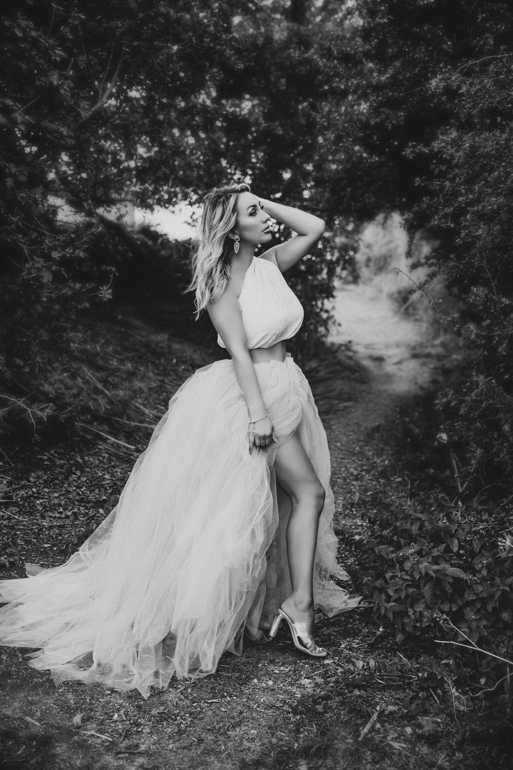 Woman in flowing white dress poses dramatically on a forest path in black and white.