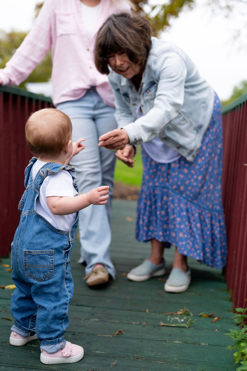 A toddler in denim overalls interacts with others on a wooden deck during a fall day.