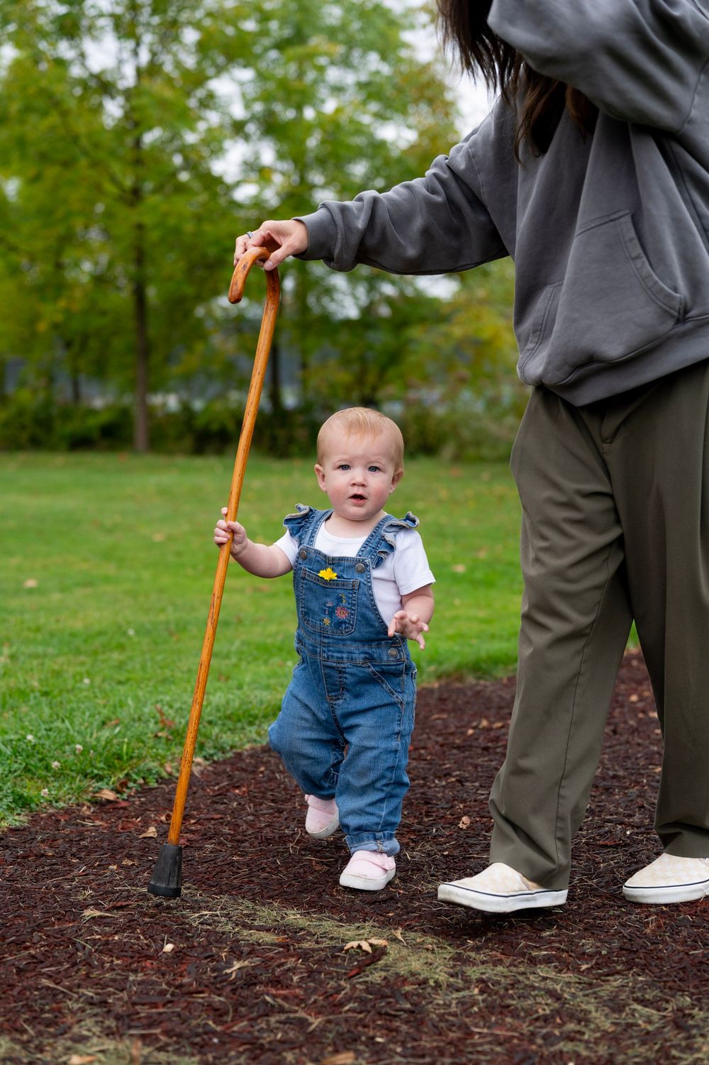 A small child in denim overalls walks with support on mulch-covered ground next to an adult with a wooden cane.