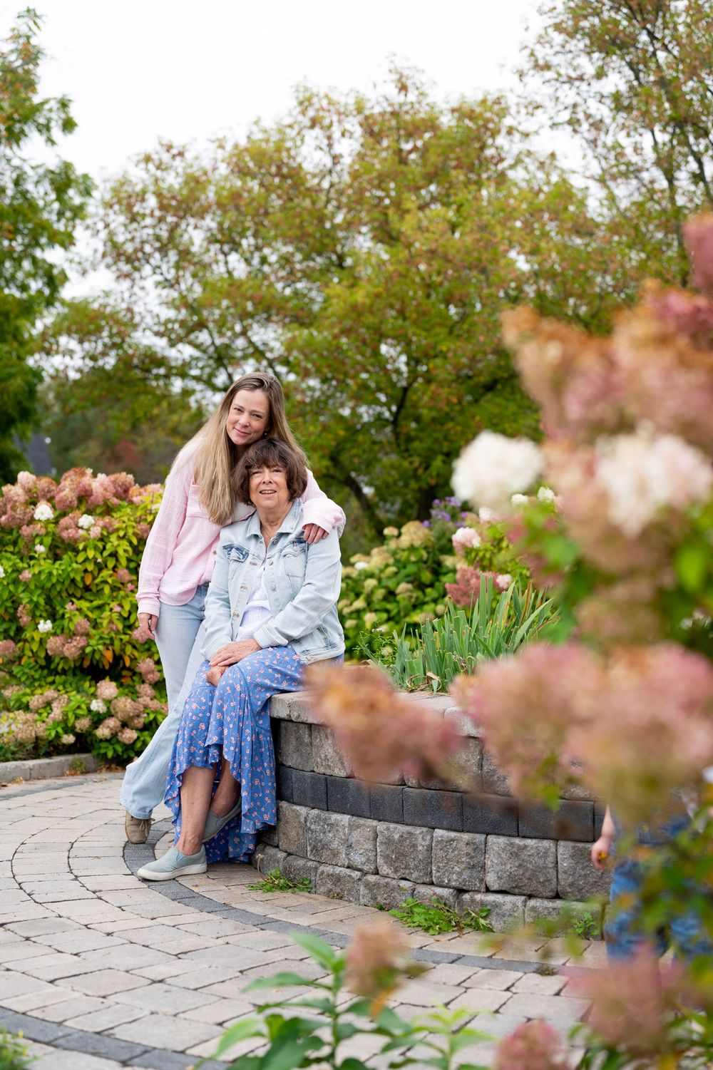 Two people sit together on a stone wall in a garden with blooming pink flowers and greenery.