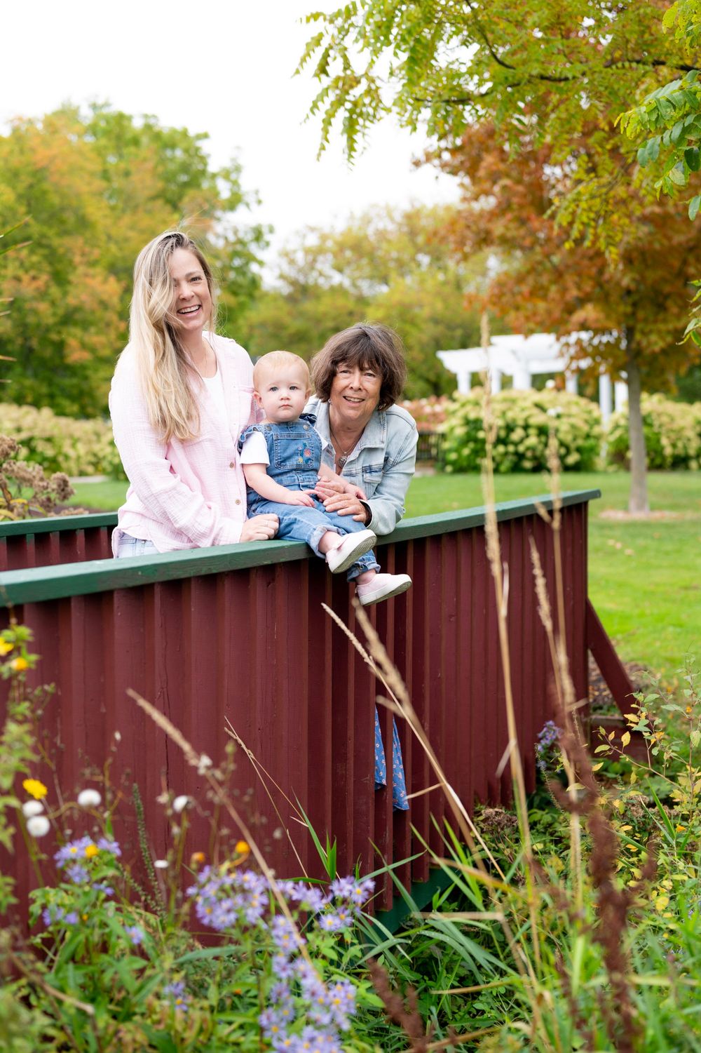 Three people enjoying a peaceful moment together on a wooden railing in a garden with blooming flowers and autumn trees.