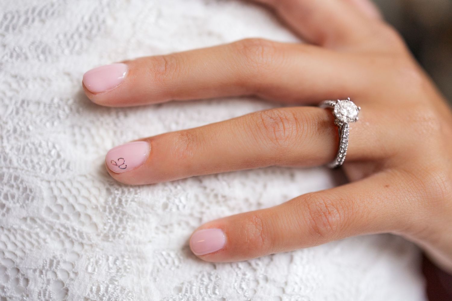 Close up of a sparkling diamond engagement ring on a hand resting against white lace fabric.