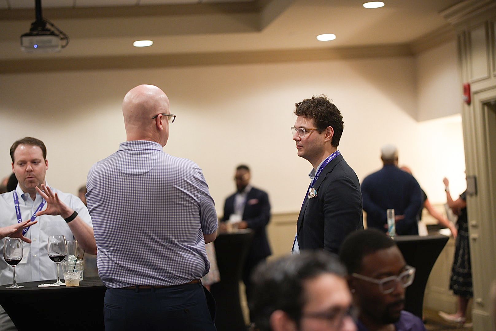 Conference attendees in business attire chat in small groups near high-top tables at a networking reception in a banquet room.