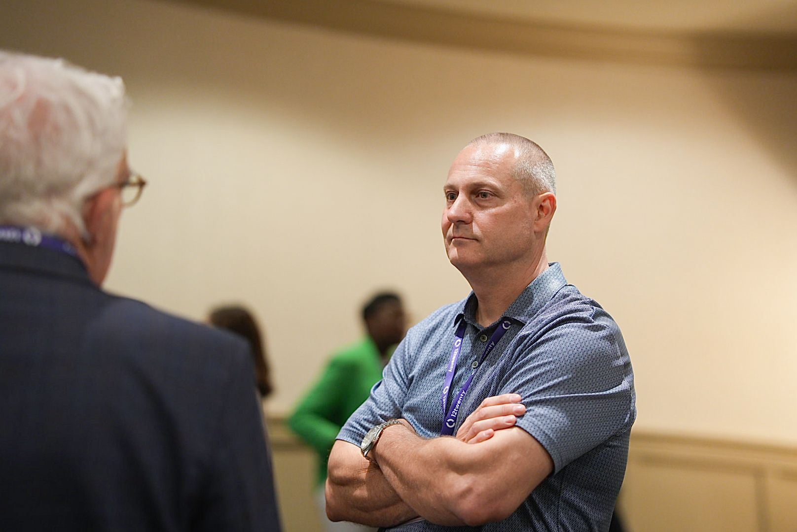 Man in a gray polo with a lanyard stands with arms crossed, listening to a conversation at a professional conference event.