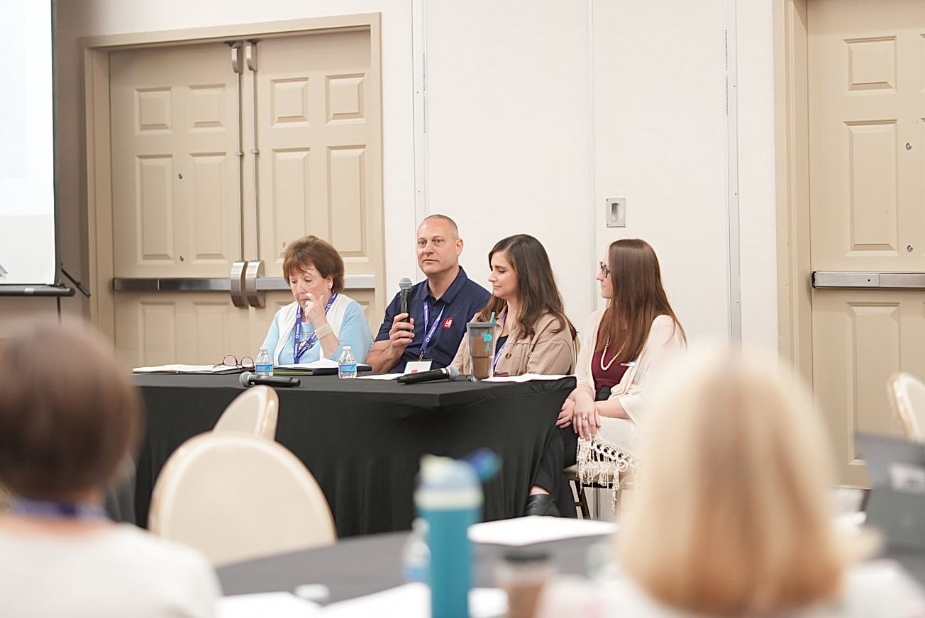 Panel of four speakers sits at a draped table facing an audience during a conference session in a meeting room.
