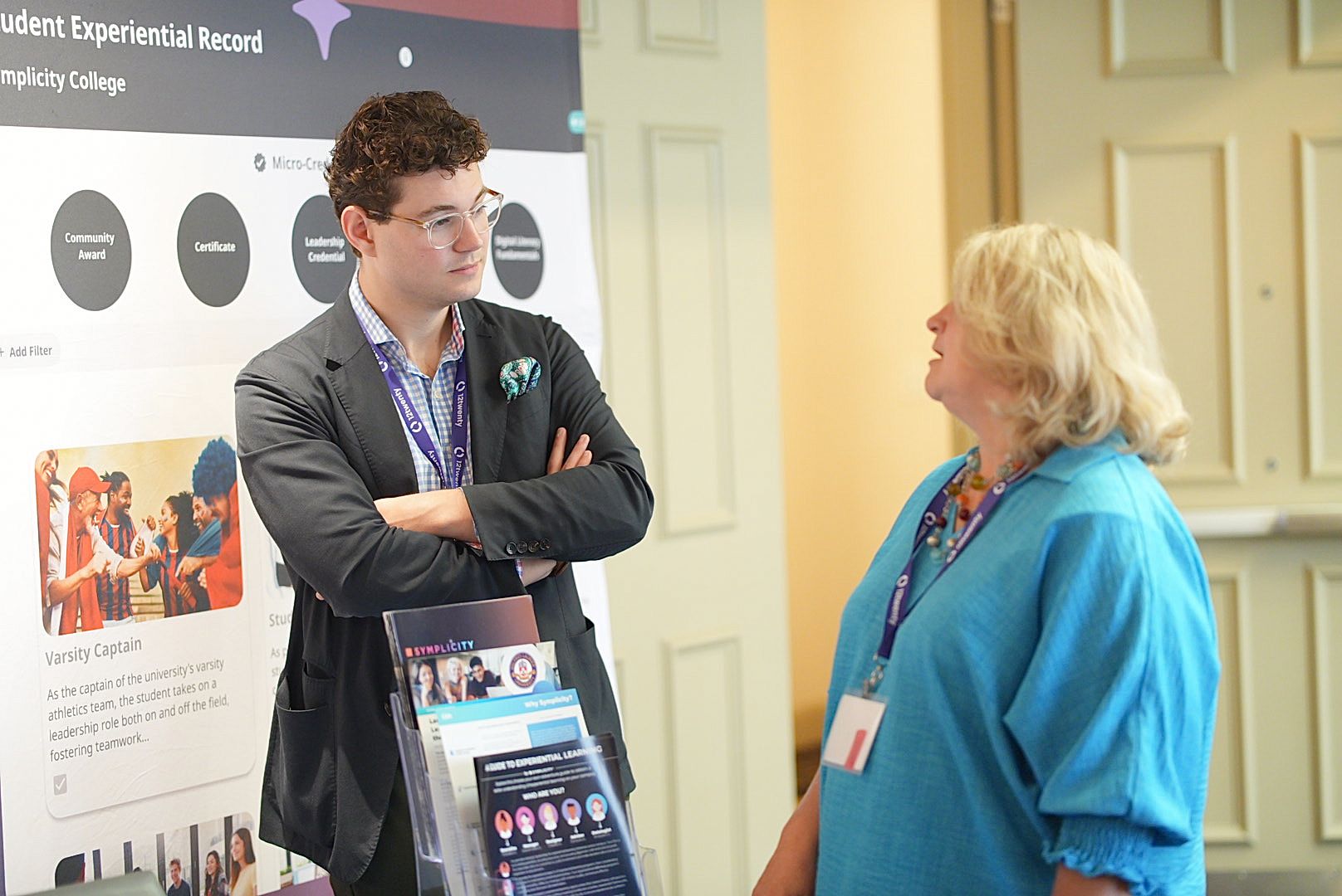 Man with glasses and a lanyard talks with a woman in a teal top beside a student experiential record display at a conference.