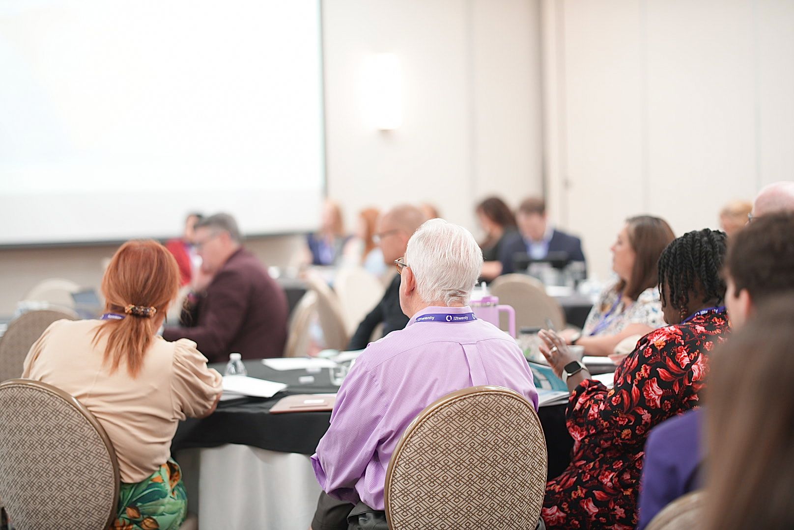 Attendees seated at round tables listen to a presentation during a conference breakout session in a bright meeting room.