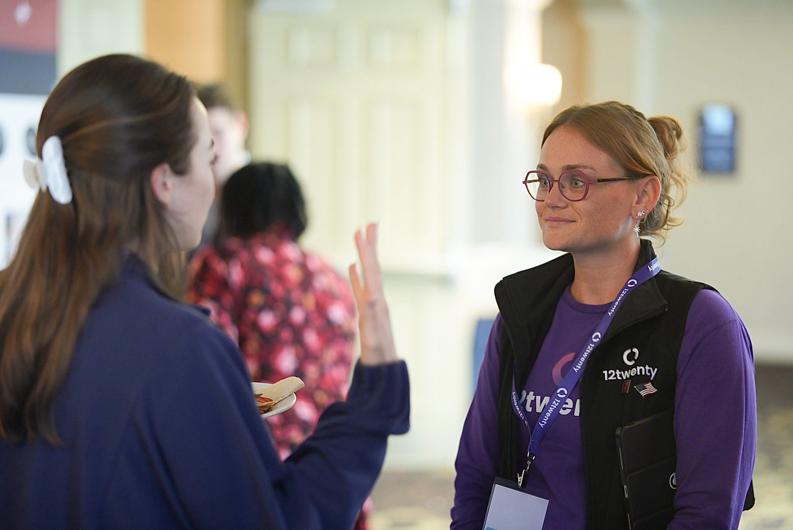 Two women in conversation at a conference, one wearing glasses, a black vest and a purple shirt with a lanyard.