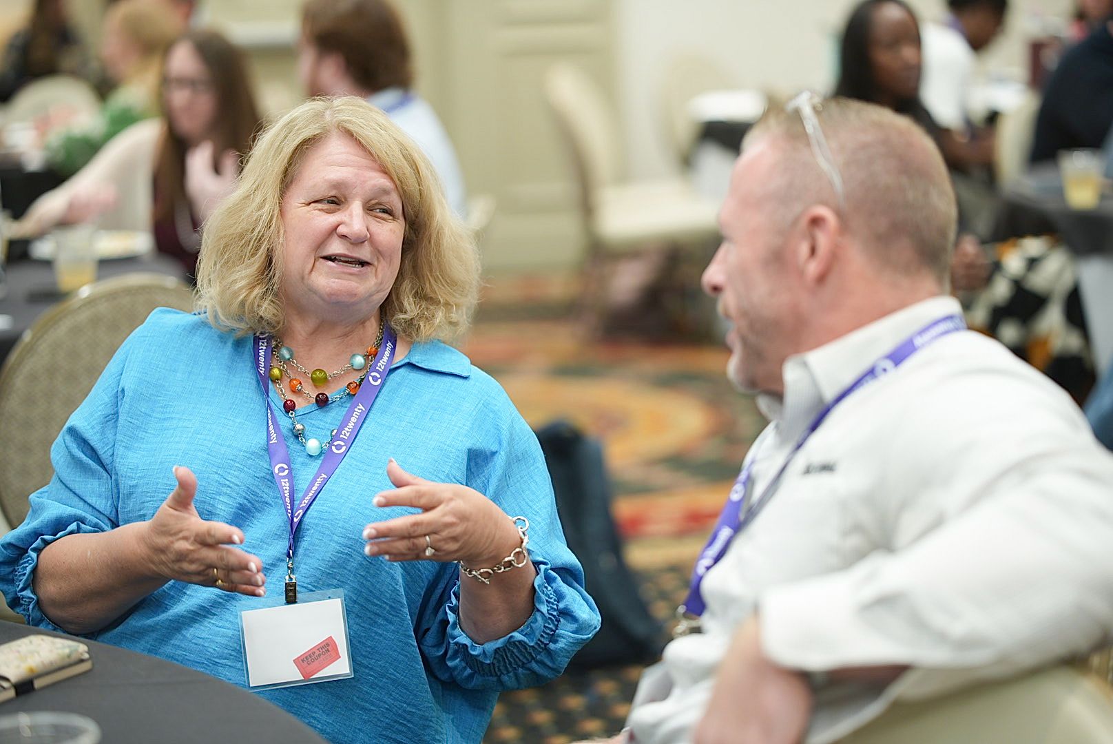 A woman in a bright blue top gestures while speaking to a man beside her at a conference table, both wearing lanyards.